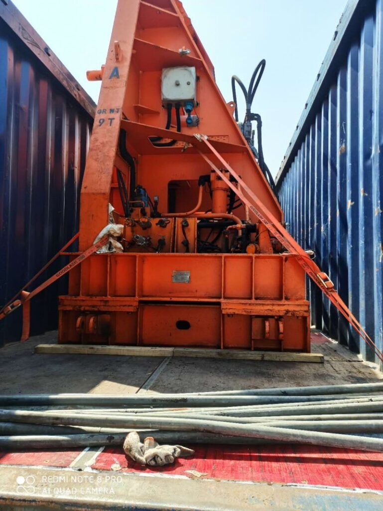 Orange hydraulic drilling rig secured on wooden skid base between two shipping containers with red lashing straps
