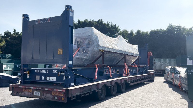 Large industrial equipment wrapped in plastic film and secured with orange straps inside open top container on flatbed trailer
