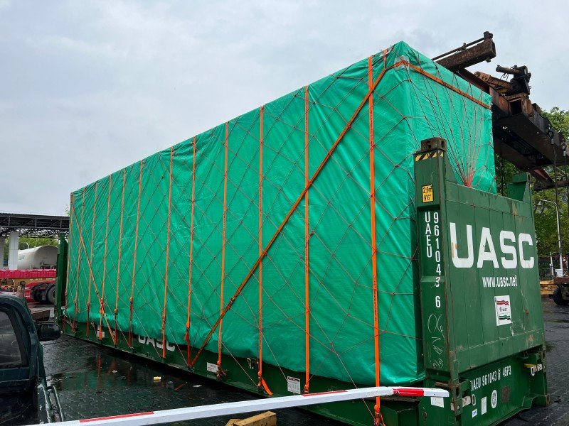 Large cargo covered with green tarpaulin and secured with diagonal orange straps on a UASC open top container