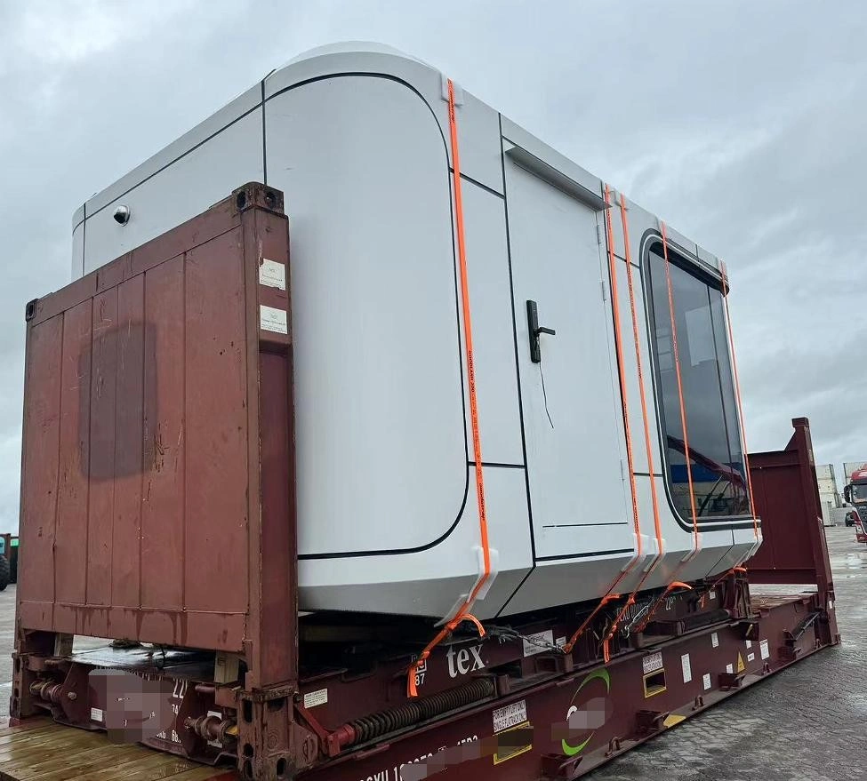 The side perspective of a white modular cabin unit loaded onto a flat rack container, showing vertical orange straps anchored to the base and structural framing used to maintain load integrity during transit.