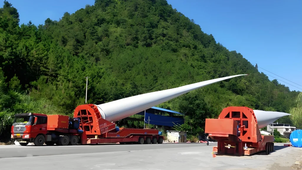 A red specialized transport trailer with a self-propelled modular transporter (SPMT) configuration carries a long white wind turbine blade through a mountainous region.