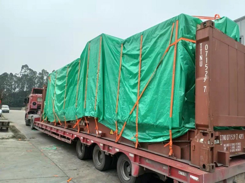 Green-tarp-covered heavy equipment on TINU open top container + flatbed; weatherproof & lashed for cross-border shipment.