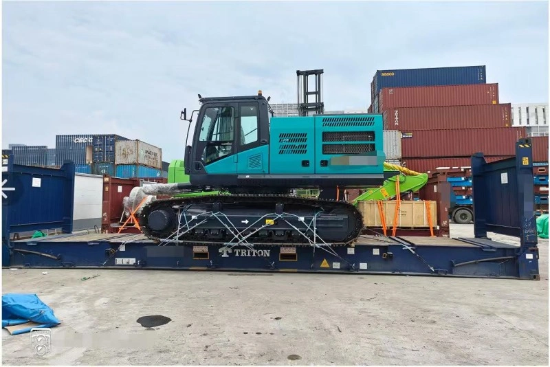 A teal-colored excavator is firmly secured with chains and straps on a blue Triton-branded flat rack container, with a forklift and stacked shipping containers visible in the background.