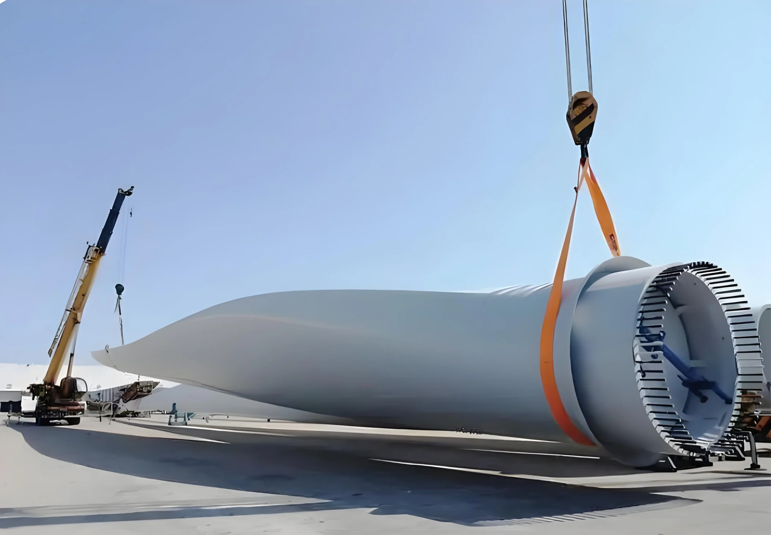 A large white wind turbine blade is being lifted by a crane using orange heavy-duty straps, showcasing the complex handling required for oversized renewable energy components.