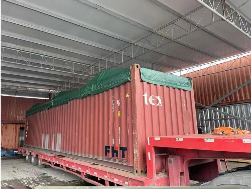 A red open top shipping container with a green tarpaulin cover, loaded on a red flatbed trailer inside a warehouse, ready for heavy cargo transportation.