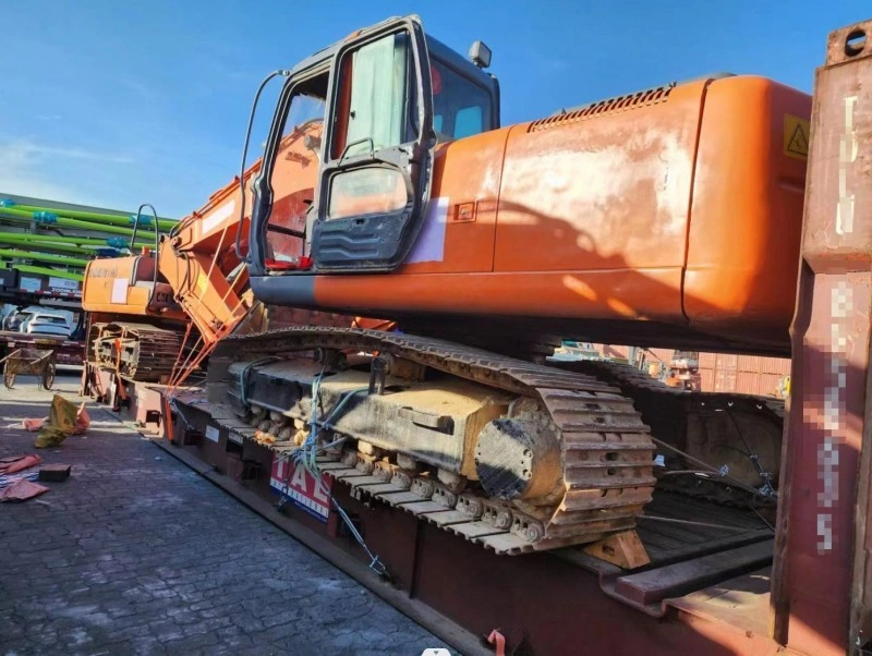 A weathered orange crawler excavator is positioned on a rusty red flat rack container, with its tracks chained down and cabin door open, ready for international shipping.