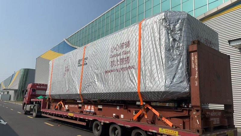 Large industrial equipment wrapped in silver waterproof tarpaulin and secured with orange straps on flatbed trailer for OOG shipment