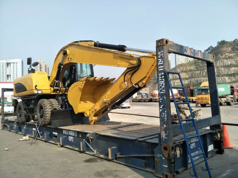Yellow wheeled excavator secured on WAN HAI framework flat rack (WHLU 4065813) at port; steel blocking & anti-slip mats applied.