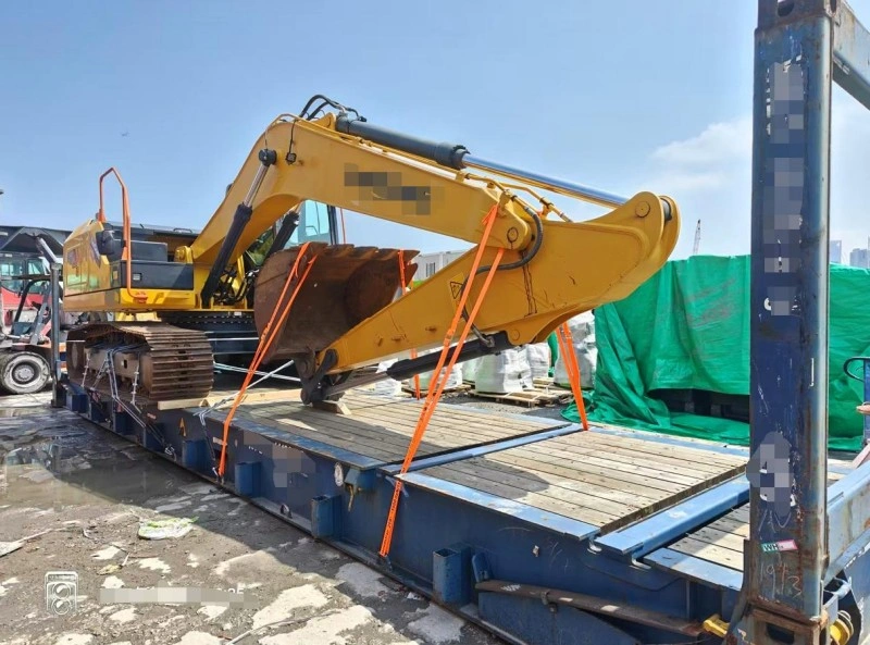 A large yellow crawler excavator is strapped down with orange tie-downs onto a blue flat rack shipping container in a port yard, ready for international transport.