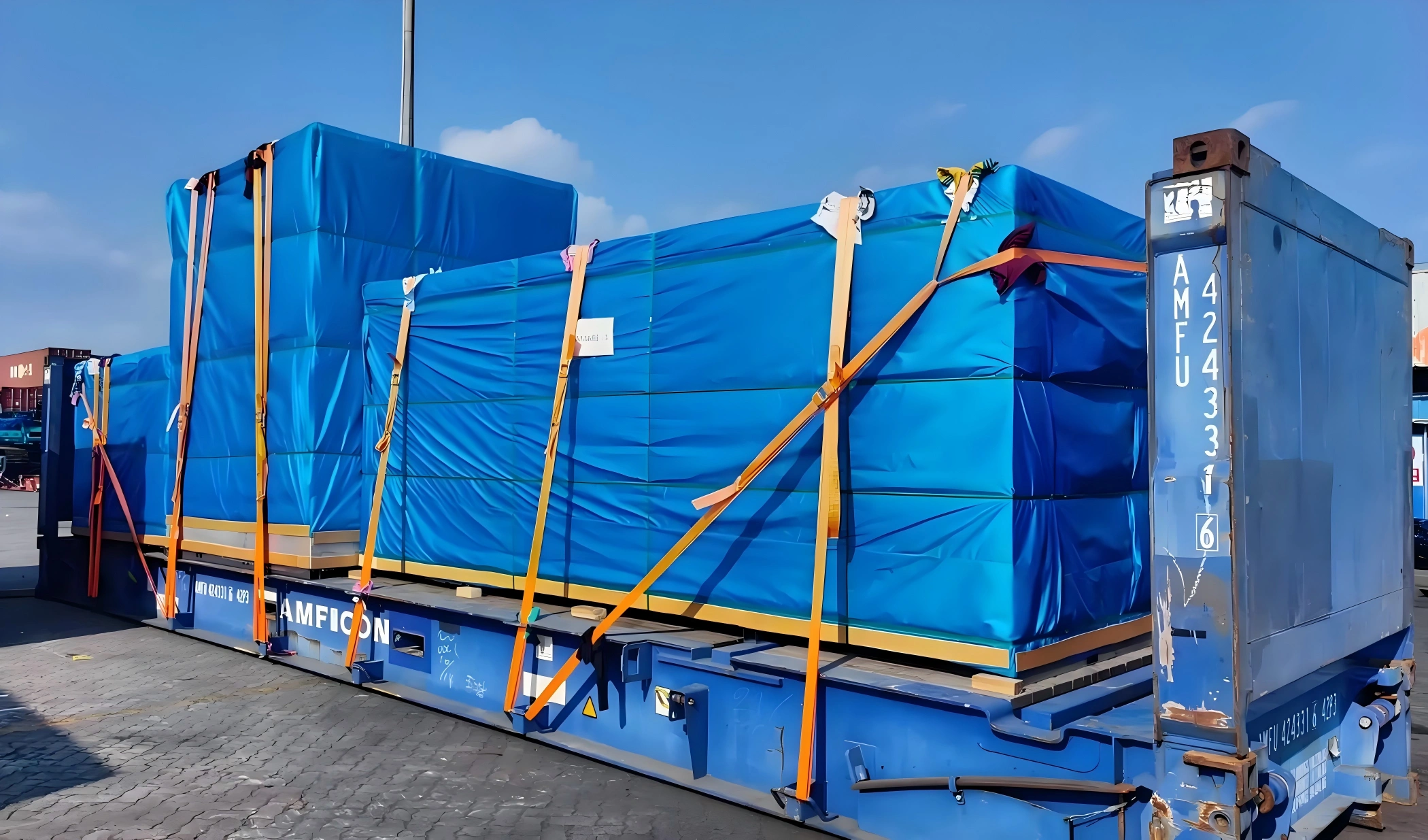 Several vacuum-prepared cargo units covered in blue tarp and lashed diagonally on AMFICON flat rack trailer for OOG export shipment