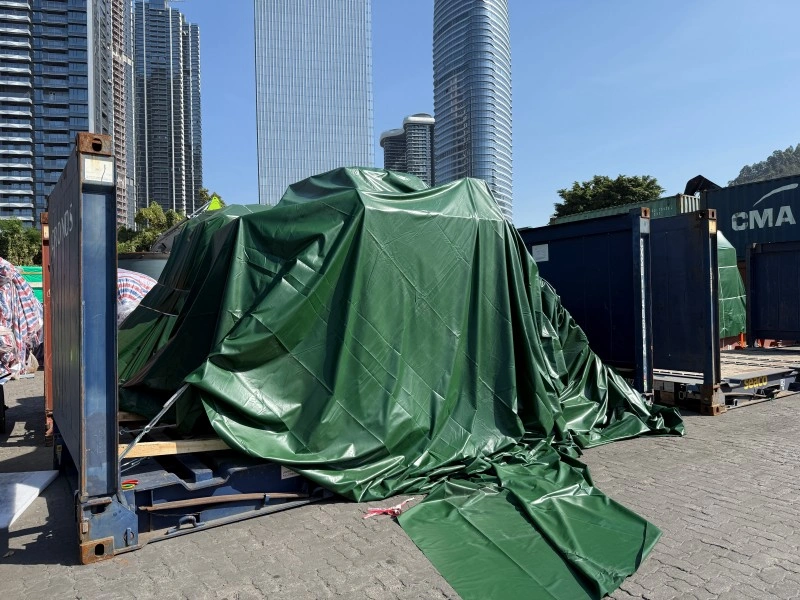 An oversized shipment on a flat rack is concealed under a heavy-duty green tarpaulin, serving as a security measure to prevent unauthorized access and theft of components during storage.