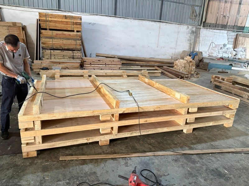 A worker uses a power drill to assemble a heavy-duty, double-decker timber pallet base in a workshop, surrounded by raw lumber and other packaging materials for custom export crates.