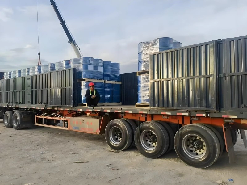 A worker in a red hard hat supervising the crane-assisted loading of blue drums onto a flatbed trailer, with stacked cargo secured for transport, highlighting the integration of real-time monitoring in heavy logistics.