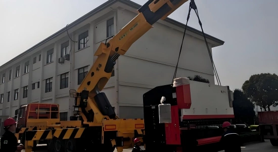 Yellow crane lifting white industrial cabinet to elevated position at factory building with workers guiding for precise placement