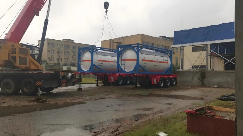 A large red and yellow mobile crane is lifting a blue-framed ISO tank container, which holds two white cylindrical tanks with orange hazard stripes. The container is being positioned onto a red multi-axle flatbed trailer in an industrial yard with buildings in the background.