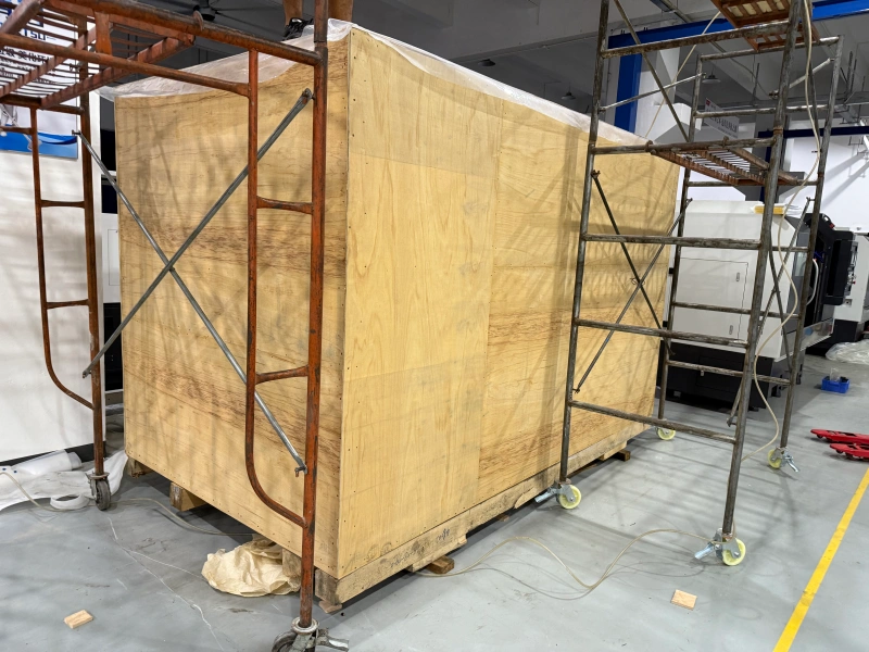 A large, reinforced plywood crate constructed around a piece of industrial equipment, surrounded by scaffolding in a warehouse facility.