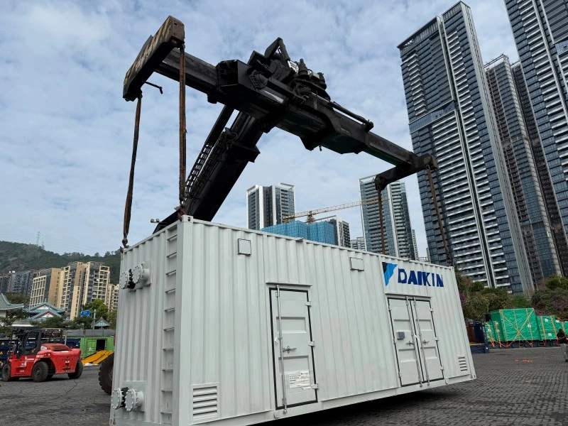 A white Daikin container being lifted by a large crane in a logistics yard, with high-rise buildings in the background.