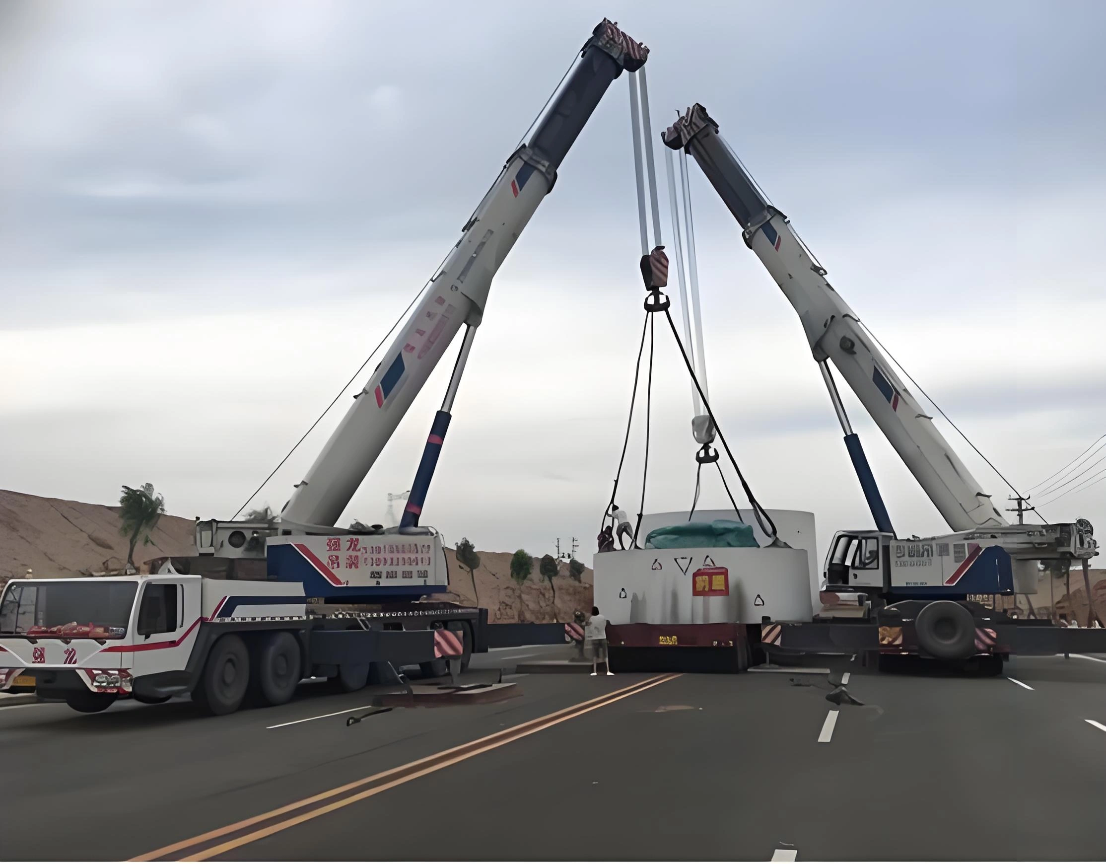 Dual mobile cranes lifting white mining equipment module during roadside installation with workers guiding for precise placement at remote site