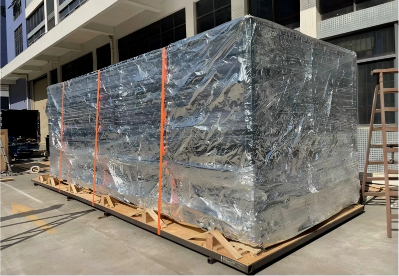 Oversized industrial unit on skid protected by dual-layer silver foil barrier and clear stretch film with orange lashing straps for flat rack transport