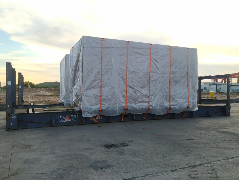 A large, shrink-wrapped industrial machine secured with orange straps on a blue flat rack container, ready for overseas transport, demonstrating precise load planning for oversized cargo.