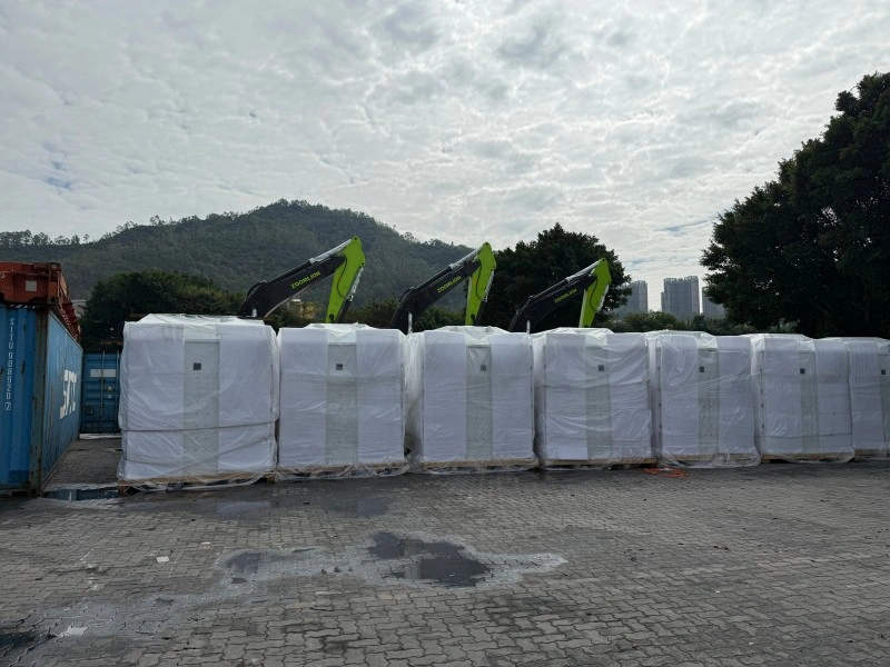 A row of new excavators, wrapped in protective white plastic, are lined up in a shipping yard with their green arms visible, ready for delivery.