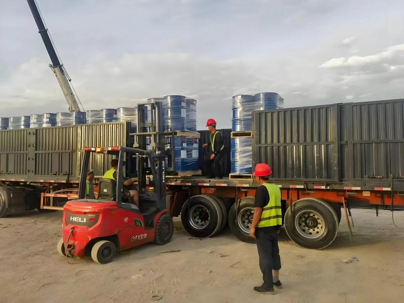 A red HELI forklift loading shrink-wrapped blue drums onto a flatbed trailer, with workers in safety vests supervising the operation under a crane, illustrating real-time coordination in oversized cargo transport.