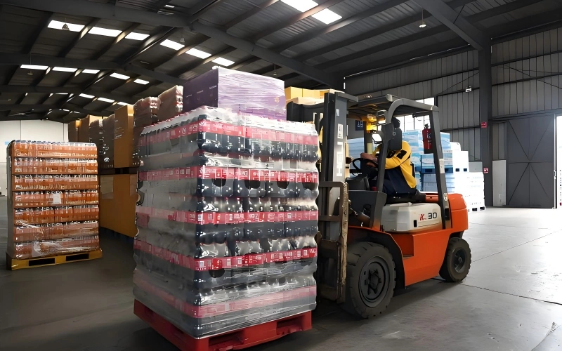Orange K 30 forklift transporting shrink wrapped pallet of bottled beverages inside temperature controlled warehouse with additional pallets staged nearby