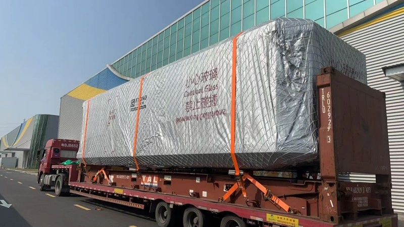 A large glass product wrapped in silver protective material with "Caution Glass" warnings is transported on a flatbed trailer, showcasing specialized handling for fragile OOG shipments.