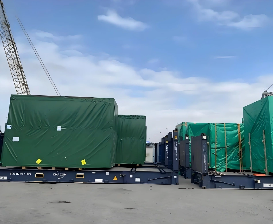 Green tarp covered industrial modules loaded on CMA CGM flat racks at port terminal with crane overhead for export shipment staging