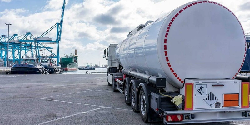 White hazardous material tanker truck parked at port terminal with shipping containers and cranes in background, displaying danger placards