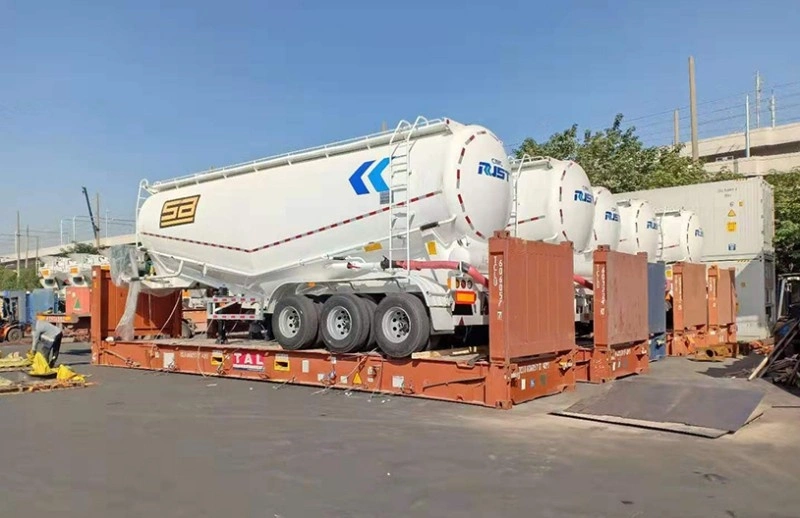 A large, white bulk cement tanker trailer with "RUST" branding is securely loaded onto a red flat rack container in an outdoor logistics yard, with other shipping containers visible in the background.