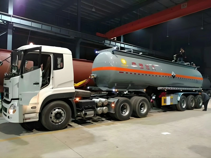 A white heavy-duty truck tractor connected to a large, grey cylindrical tank semi-trailer inside a warehouse. The tanker features an orange horizontal stripe and Chinese characters indicating it is designed for transporting corrosive liquids, with workers inspecting the vehicle.