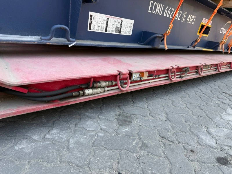 A close-up view of a blue flat rack container with the ID ECMU 662 62 45P3, secured by orange lashing straps on a red trailer bed, ready for transporting oversized cargo.