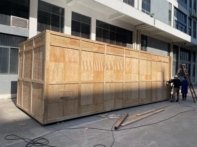 A massive wooden export crate being assembled by workers outside a factory, designed to protect industrial machinery during international shipping.