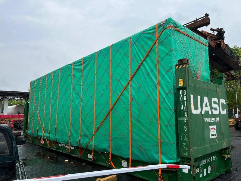 A large cargo unit wrapped in green tarpaulin and secured with a net of orange straps is being lifted by a crane towards a green UASC flat rack container, highlighting heavy lift logistics.