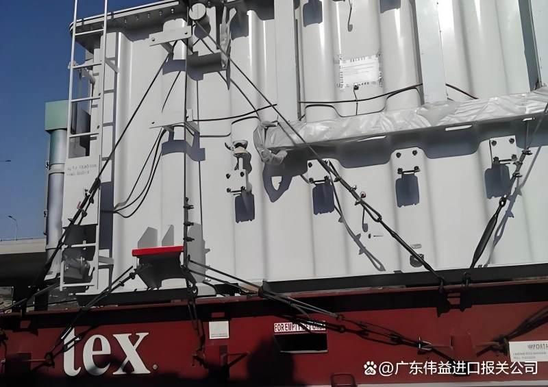 A close-up view of a large, white piece of heavy machinery secured to a red flat rack container with multiple black lashing straps and turnbuckles, highlighting the robust securing points and protective wrapping.