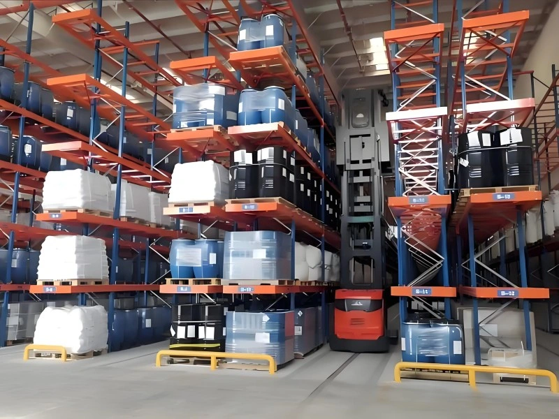 A high-bay warehouse with orange and blue pallet racking systems storing numerous blue and black chemical drums on pallets. A red forklift is positioned between the aisles, indicating active material handling operations.