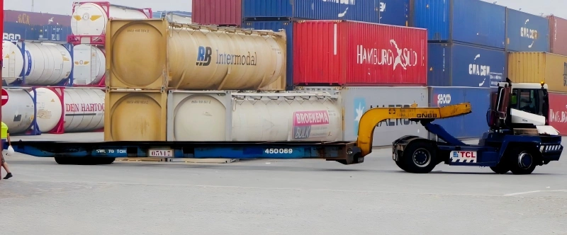 A reach stacker is moving a flat rack loaded with two stacked ISO tank containers in a busy port yard. The containers are positioned next to standard shipping containers, highlighting intermodal freight transport operations.