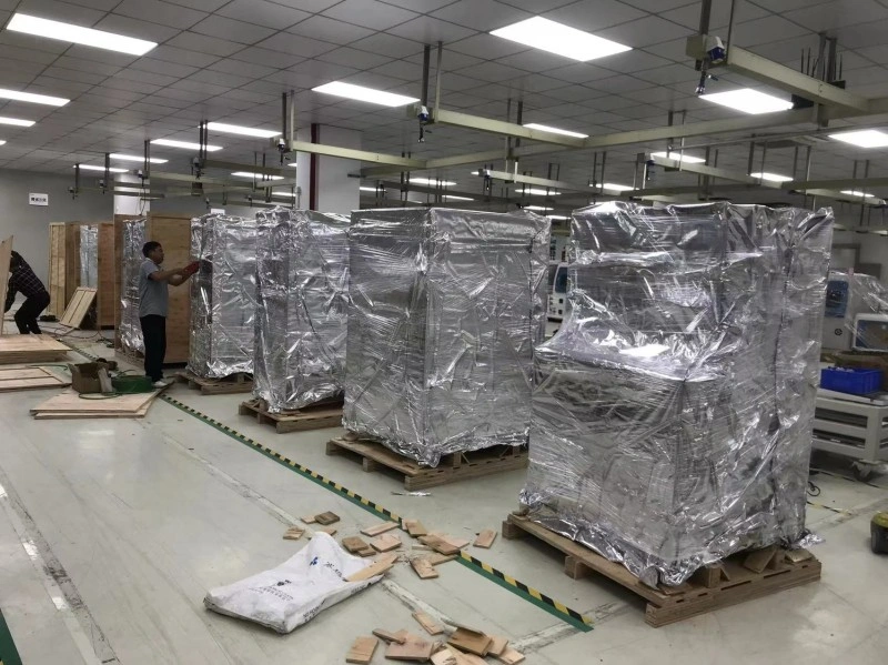 Workers in a factory facility preparing industrial machines for export, wrapping them in silver moisture-proof aluminum foil bags on wooden pallets to prevent rust and corrosion during ocean freight.