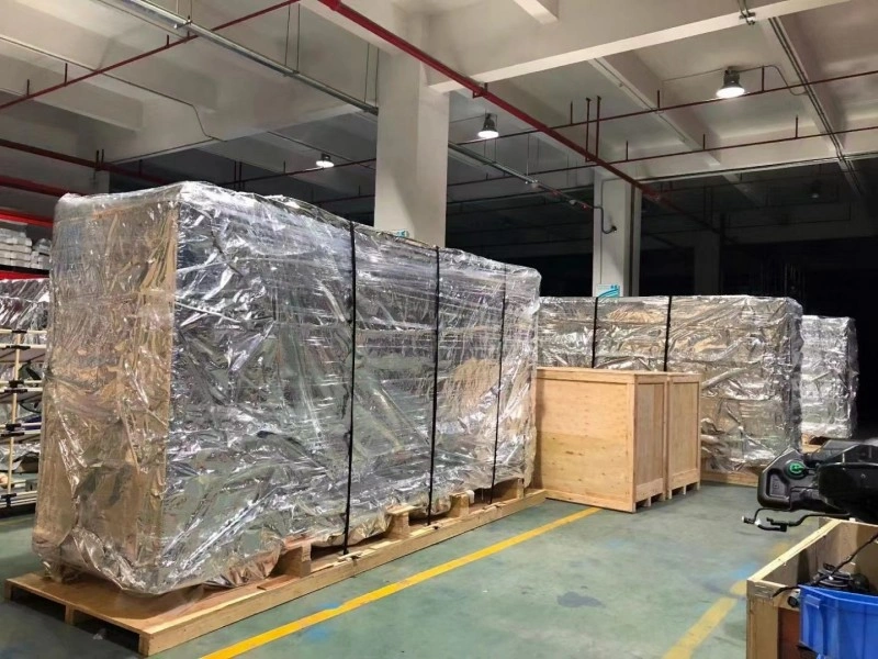 Large industrial machines on wooden pallets, fully enclosed in clear shrink-wrap and secured with black straps, stored inside a warehouse with other export-ready cargo.