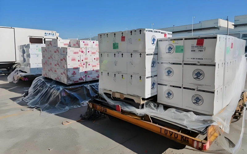 Flatbed trailer transporting stacked white insulated shipping containers with handling labels and temperature sensitive markings covered by protective plastic sheeting
