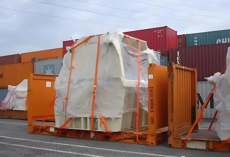 A large, white industrial machine, wrapped in protective plastic and secured with bright orange lashing straps, is loaded onto an orange flat rack container in a shipping yard, with standard shipping containers stacked in the background.