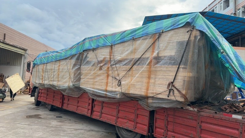 A long red flatbed truck carries a large, irregularly shaped cargo piece fully wrapped in protective plastic sheeting and covered with a blue-green tarpaulin, secured with multiple tie-down straps in an industrial yard.