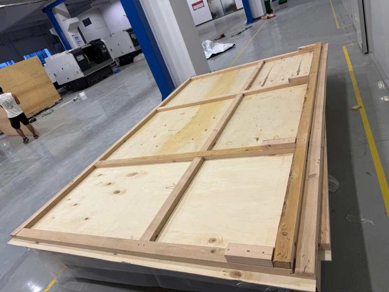 Workers are securing a large, irregularly shaped OOG cargo load on a flatbed truck using heavy-duty straps and a protective plastic tarp to shield it from weather during inland transportation.