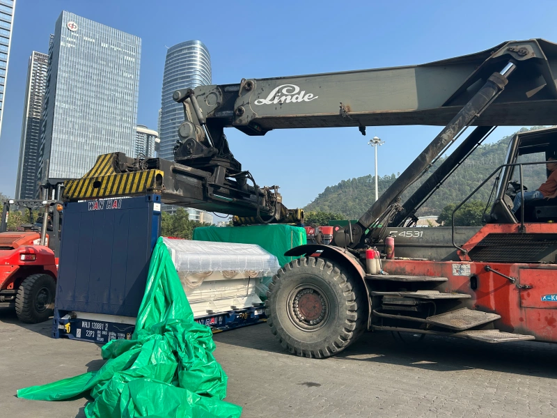 A Linde crane lifts a large, plastic-wrapped industrial component into an open top shipping container, while two workers in safety gear supervise the operation in a port yard with mountains in the background.