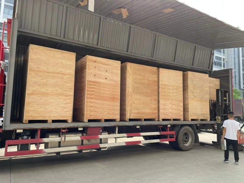 A row of large, light-brown wooden export crates on pallets being loaded onto a red flatbed truck by a forklift, supervised by a worker in a white shirt.