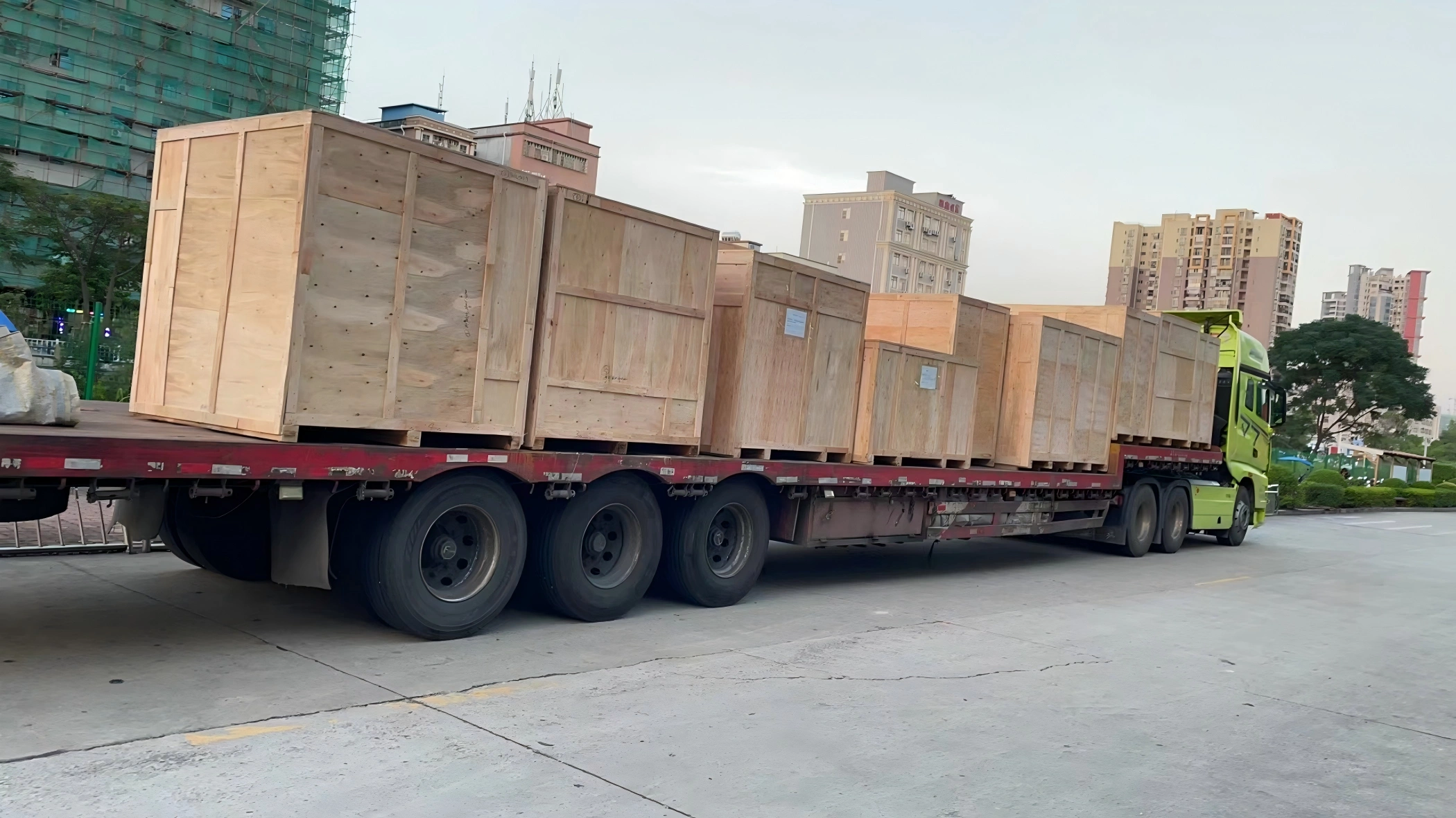 Green prime mover hauling flatbed trailer carrying six large wooden crates for oversized project cargo transport in city environment