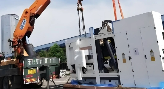 Orange mobile crane lifting white industrial machine cabinet from truck bed during on site delivery for manufacturing facility installation