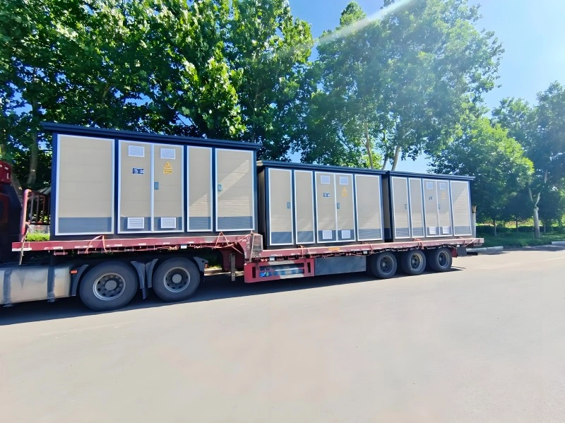 Multiple beige and blue modular power generator sets are secured on a red flatbed trailer, ready for overland transportation, with green trees in the background.