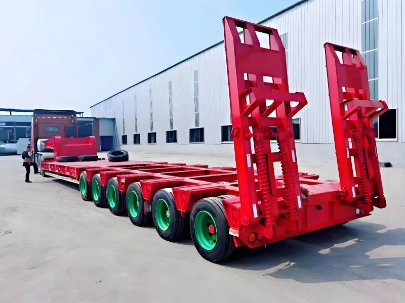 A red, multi-axle hydraulic modular trailer with green wheels is shown in a factory yard, designed for transporting ultra-heavy and oversized loads, with a worker inspecting the setup near a large industrial building.